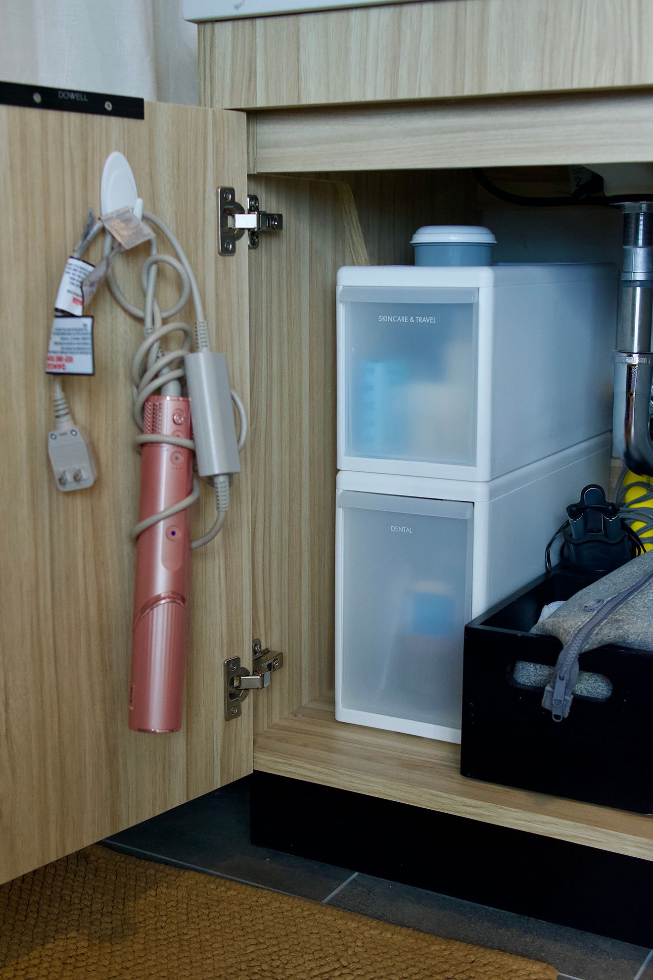 Inside a bathroom cabinet with skincare and travel storage containers, a small blue container, and a pink hair curling iron hanging on the cabinet door.