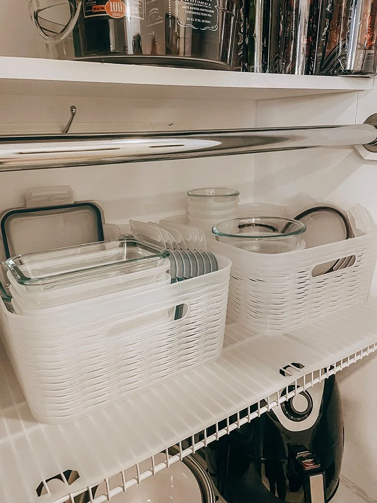 Organized kitchen shelf with white plastic baskets holding glass containers and lids, with stainless steel appliances and a black appliance below.