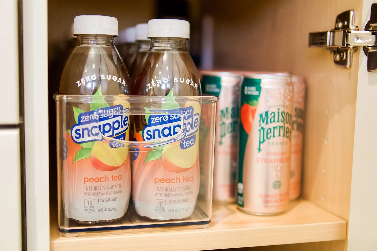 Peach flavored Snapple Zero Sugar tea bottles in a clear plastic carrier with canned strawberry-flavored Maison Perrier sparkling water behind on a wooden shelf.