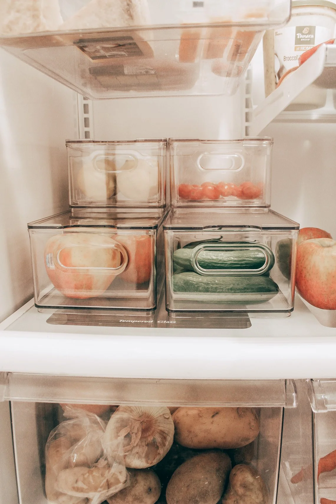 Inside a refrigerator, there are clear containers with strawberries, apples, and cucumbers, along with garlic and potatoes in the lower drawer.