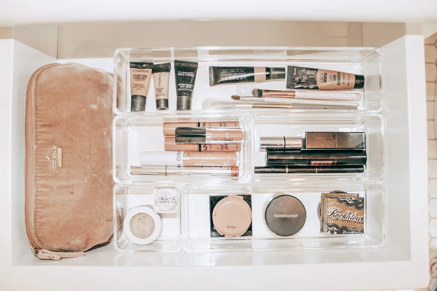 Drawer with a clear acrylic makeup organizer containing various makeup products including foundations, lip glosses, concealers, and eye shadows, with a beige velvet pouch on the left side.