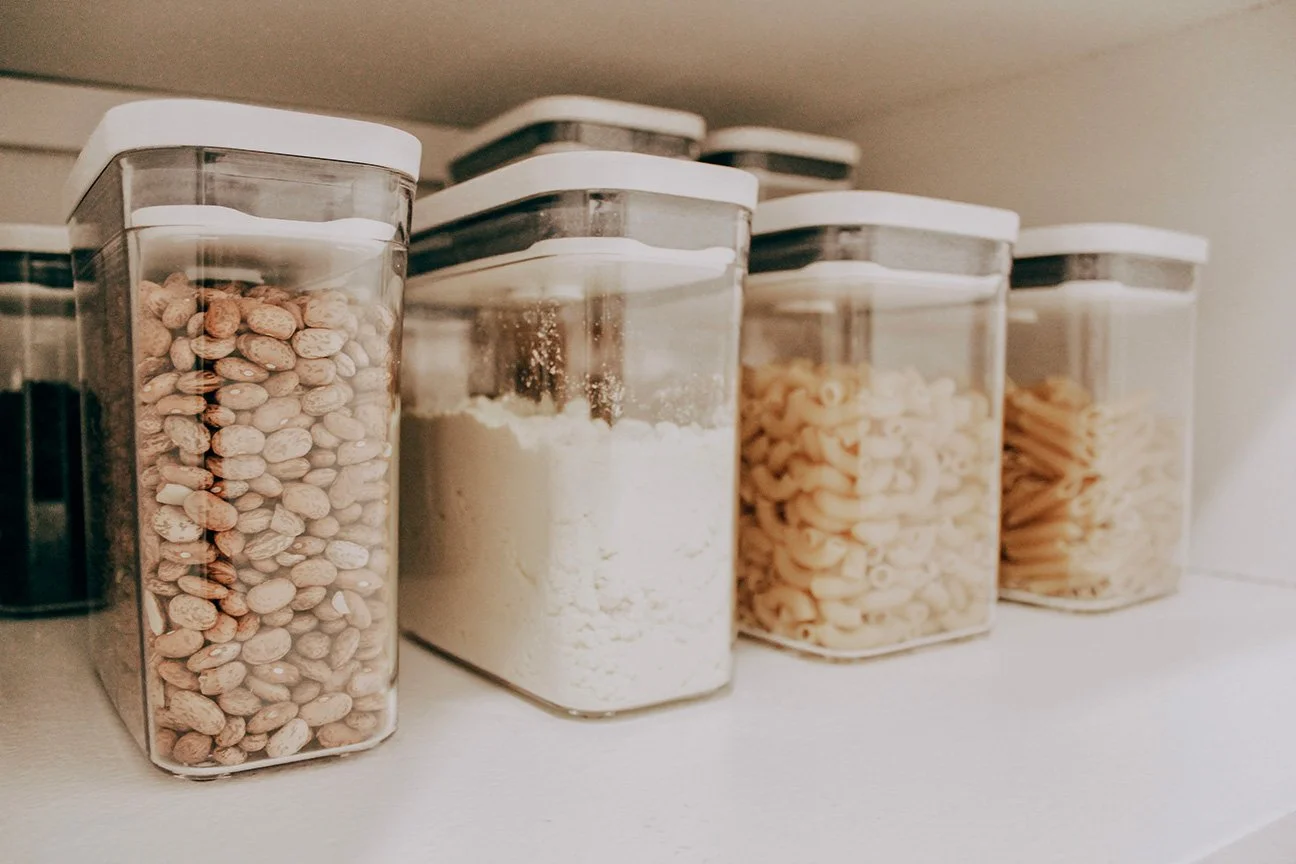 Four clear plastic storage containers with white lids, filled with different types of food staples, lined up on a white shelf.