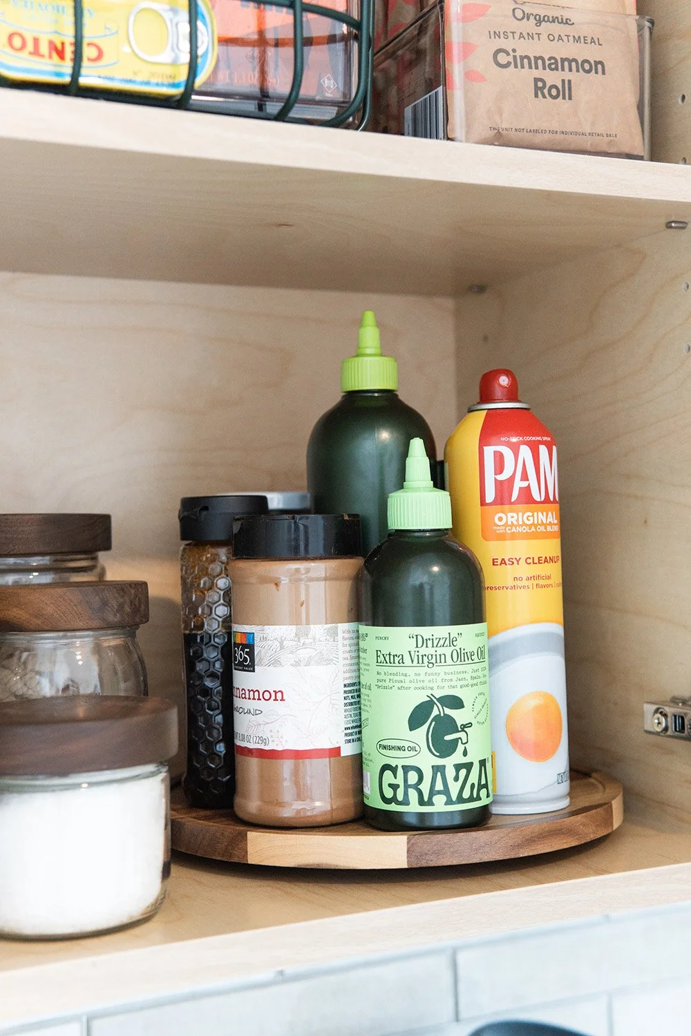 Assorted kitchen items on a round wooden shelf, including bottles of extra virgin olive oil, cinnamon, and Pam cooking spray, with jars nearby and a box of organic cinnamon roll mix on the upper shelf.