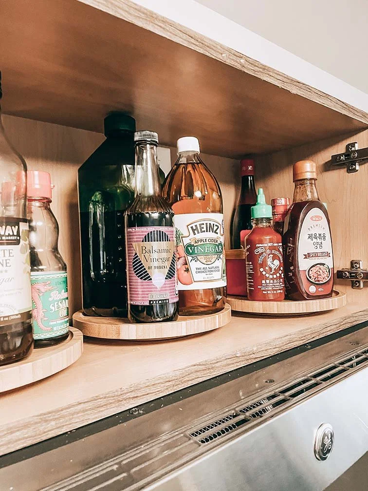 A cabinet shelf holding various bottles of condiments including vinegar, soy sauce, apple cider vinegar, hot sauce, and other sauces.