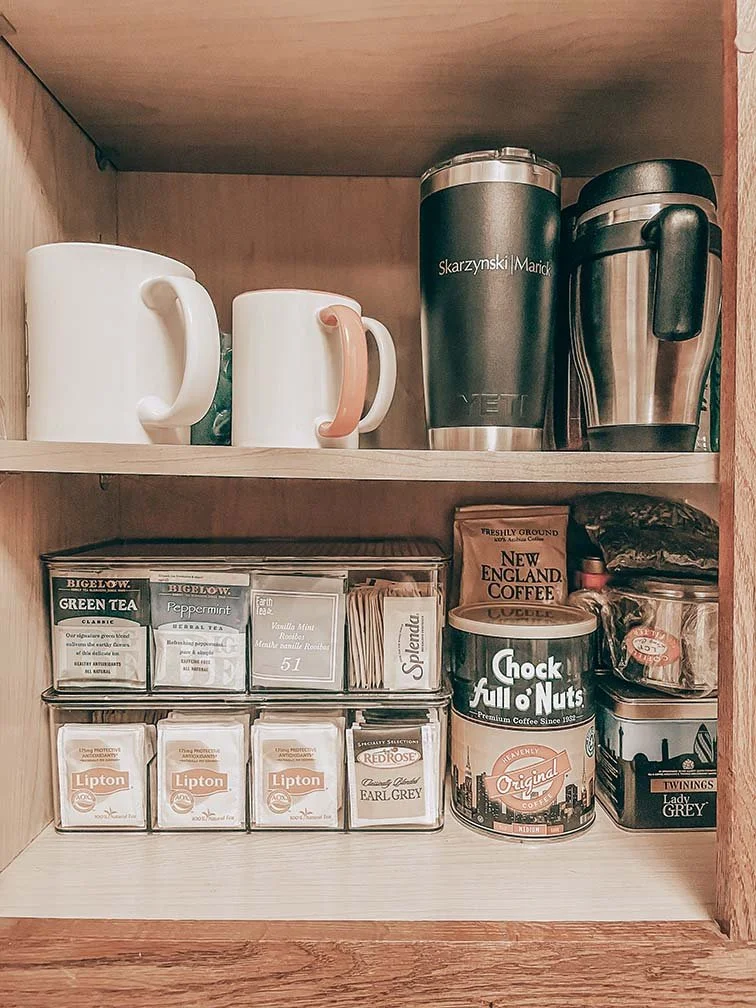 Inside a wooden kitchen cabinet, two ceramic mugs, one white and one with a pink handle, are on the top shelf. Two travel mugs, one black and one stainless steel, are next to the mugs. The lower shelf contains various tea and coffee supplies, includi