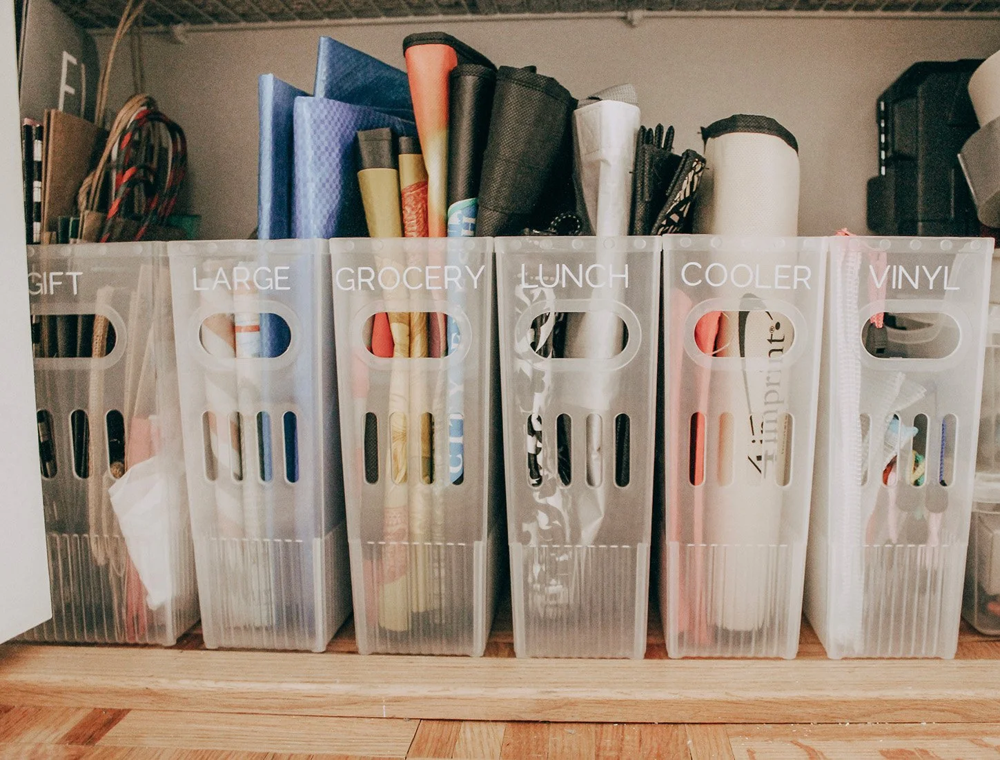 Clear plastic storage bins labeled Gift, Large, Grocery, Lunch, Cooler, Vinyl, filled with various rolled paper and crafting supplies, on a wooden surface.