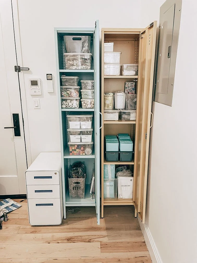 Two tall storage cabinets filled with various office supplies, paper, and organizing containers, next to a small white filing cabinet and a door.