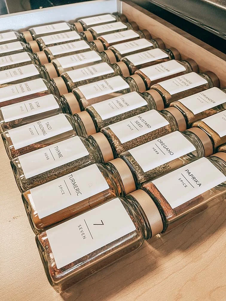 Multiple glass jars labeled with various spices such as thyme, turmeric, oregano, and cumin, arranged in a wooden spice rack drawer.
