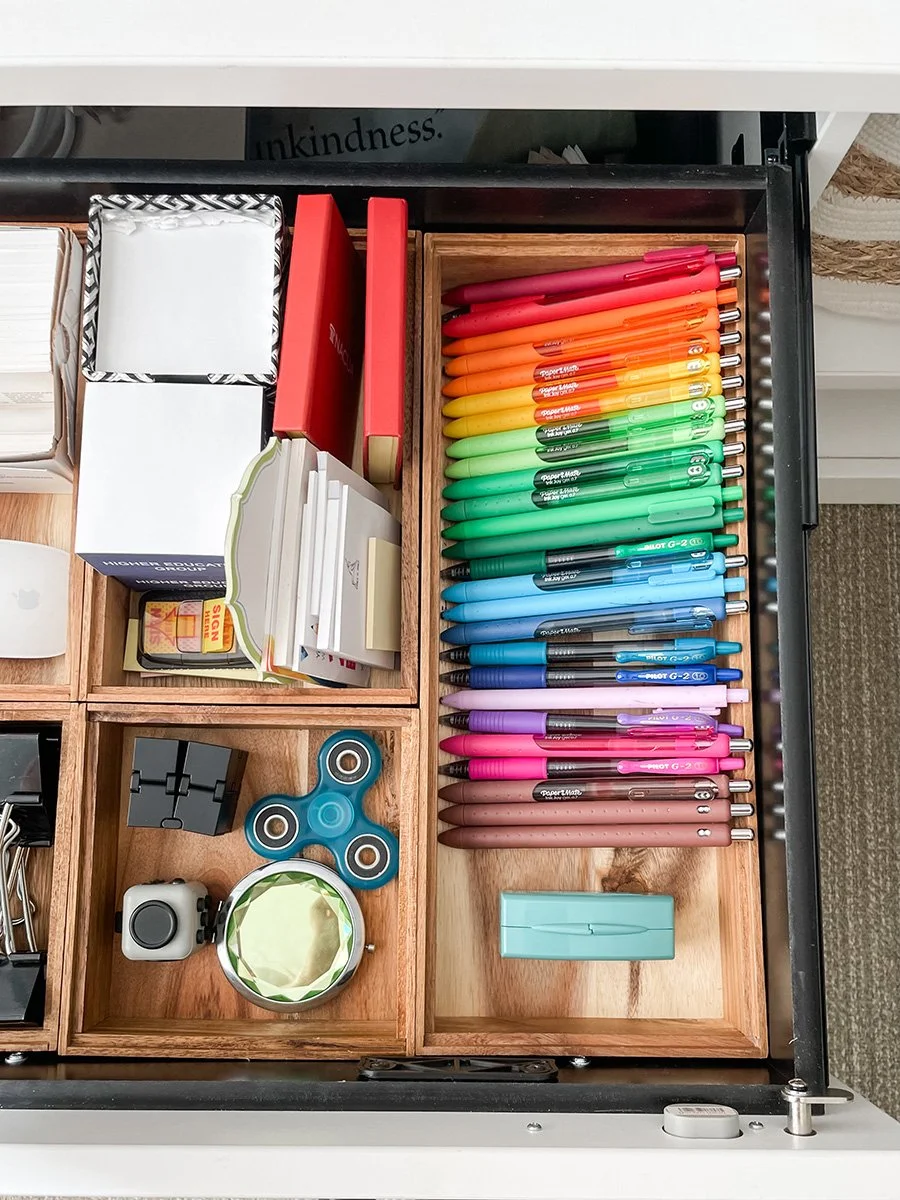 Open desk drawer containing colorful pens, a fidget spinner, a small mirror, a stapler, sticky notes, and other office supplies.