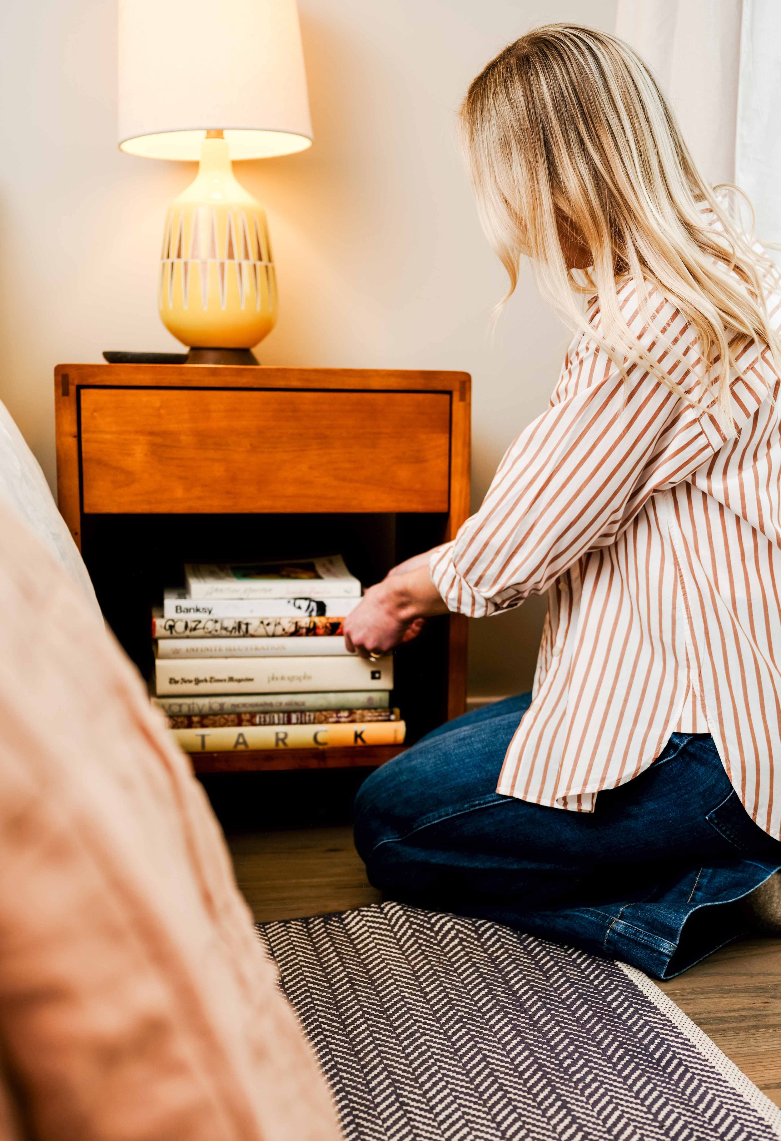 Person with long blonde hair wearing a striped shirt, kneeling on a patterned rug, organizing books on a small wooden bookshelf in a cozy room with a lamp.