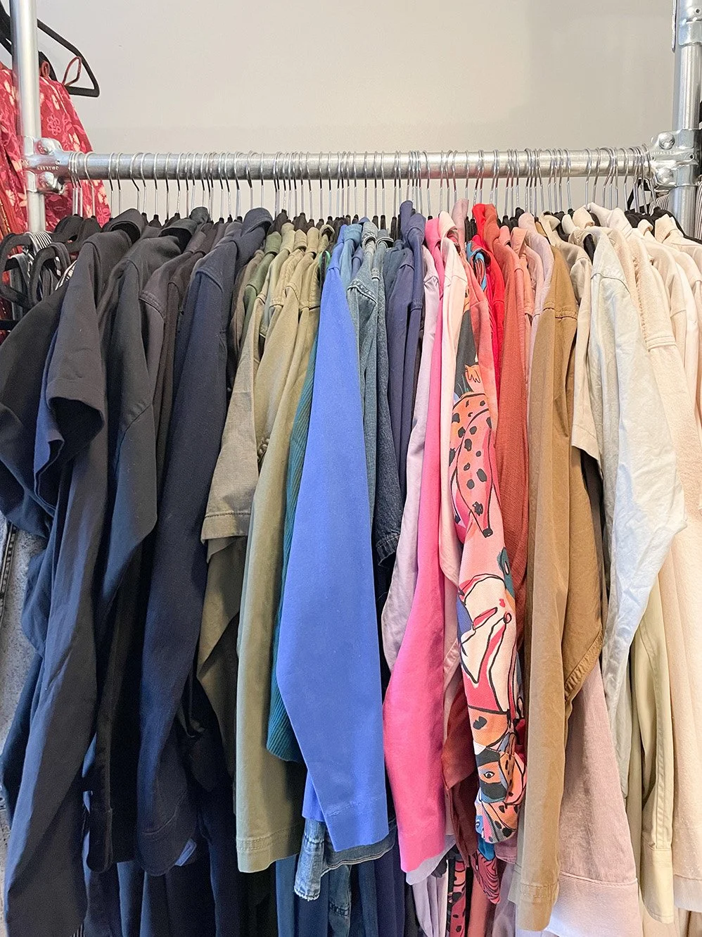 Clothes hanging on a metal rack, organized by color, including black, gray, green, blue, pink, red, brown, and beige shirts and jackets.