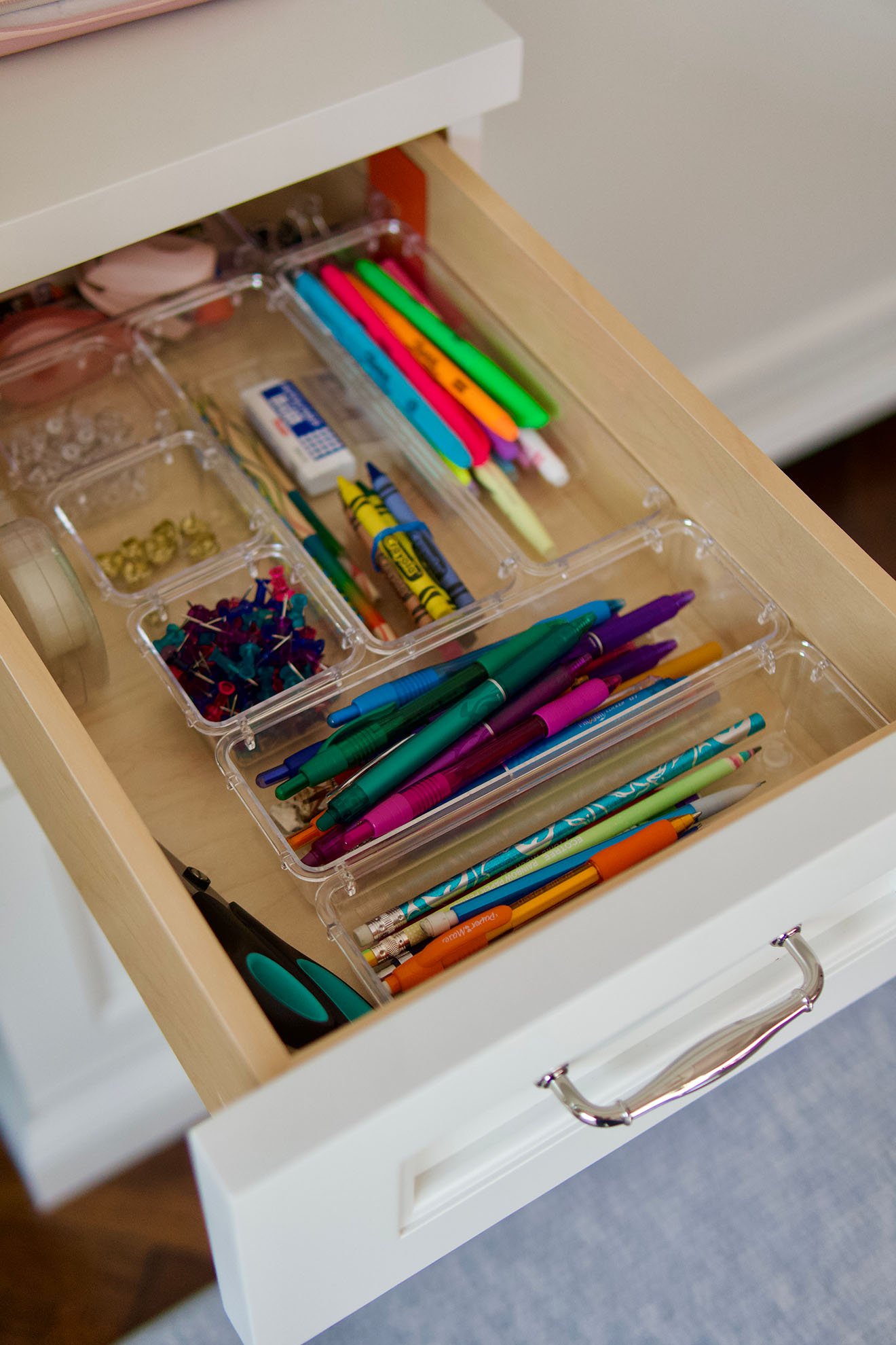 An open drawer containing various school or office supplies such as colored pens, pencils, markers, push pins, and an eraser, organized within clear plastic compartments.