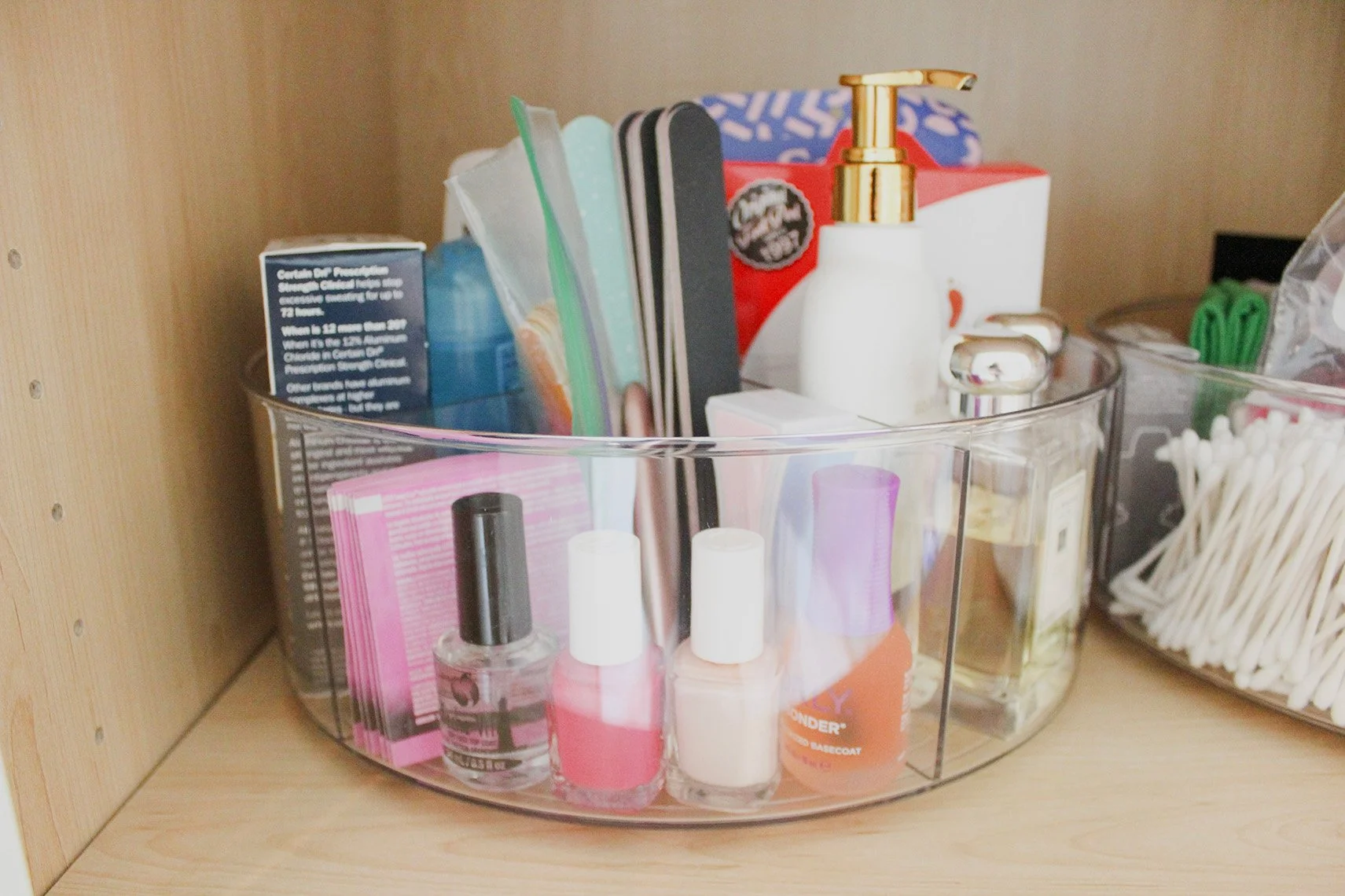 Clear plastic organizer with nail polish bottles, lip balm, and various small beauty items, placed inside a wooden cabinet.