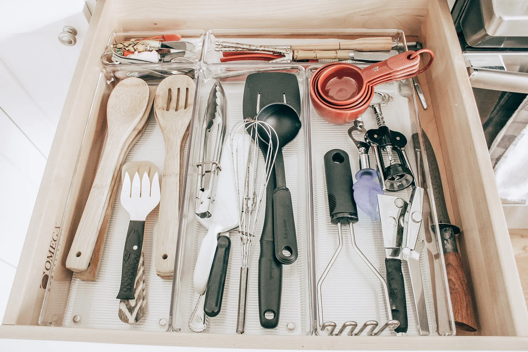 Kitchen drawer containing wooden cooking utensils, metal whisks, black and metal spatulas, small silicone spatula, plastic measuring cup, can opener, garlic press, and various kitchen tools.