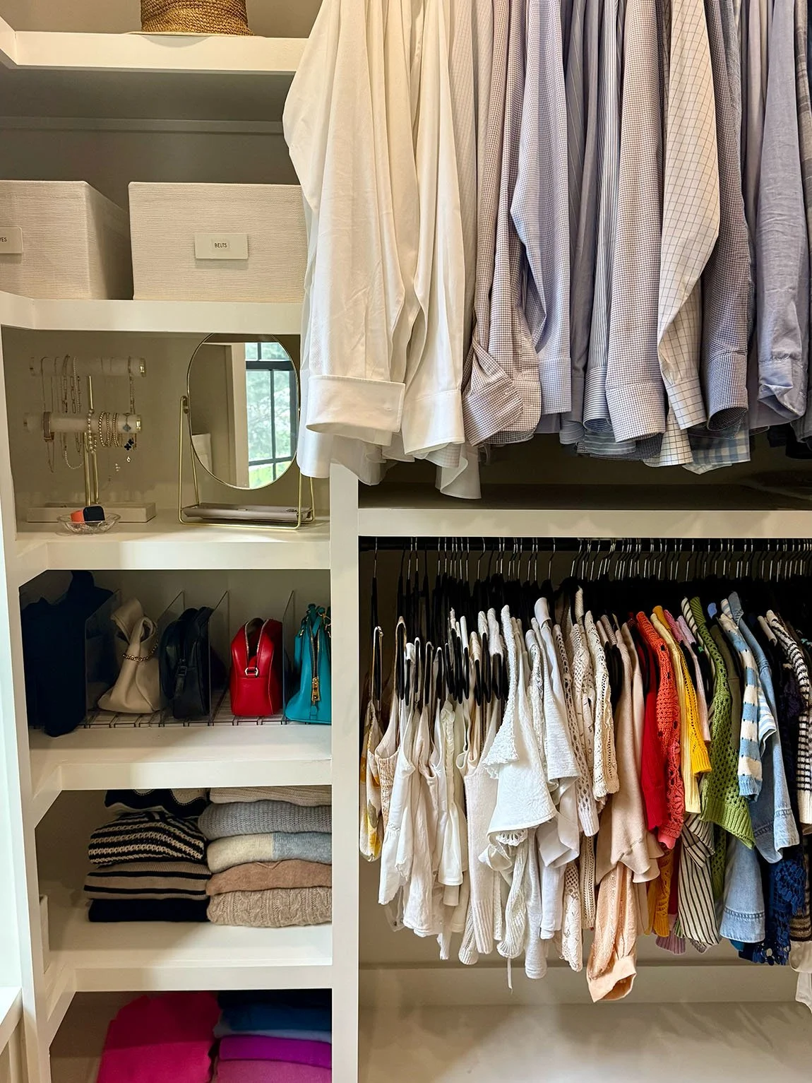 Closet with white shelving and hanging clothes, including button-up shirts, and sweaters. Shelves contain bins with labels for accessories along with purses, jewelry, and folded clothing.