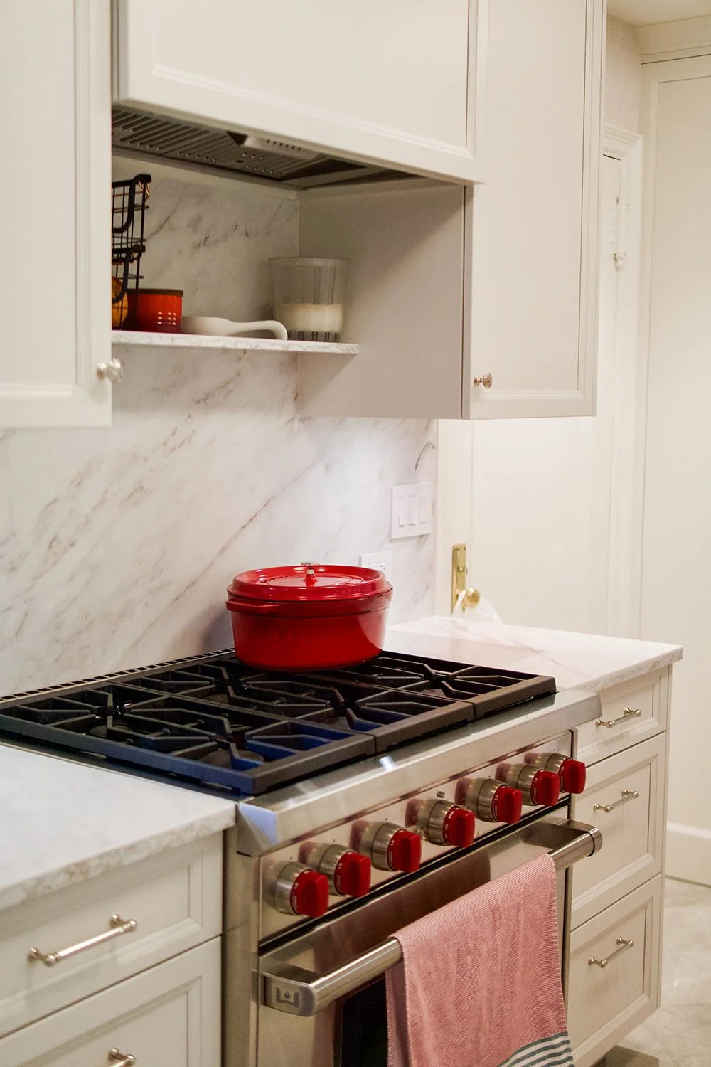Kitchen stove with a red pot on top, white cabinets, marble backsplash, and dishware on the shelf above.