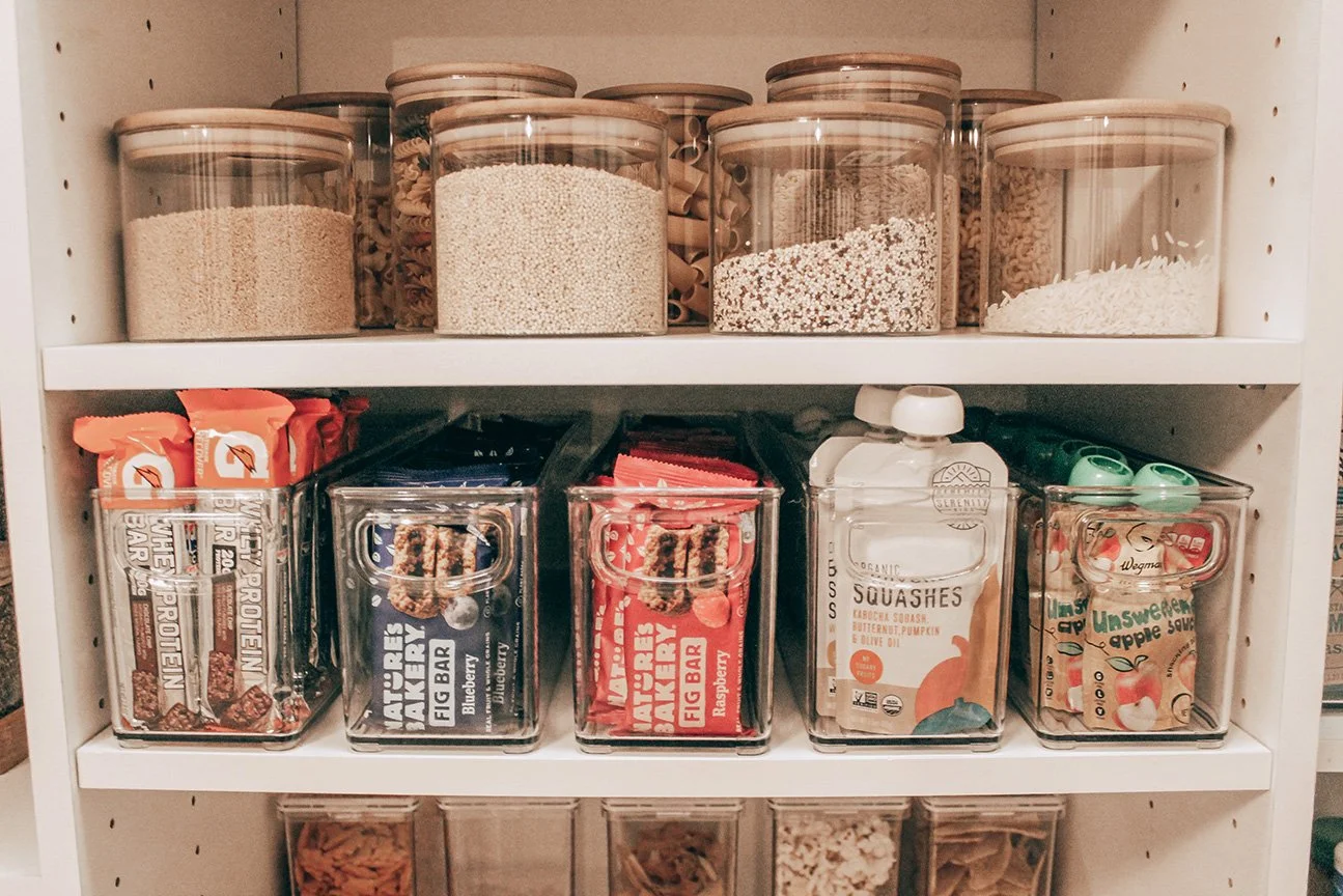 Shelf with glass jars of dry pasta and snacks, including protein bars, snack squares, and fruit snacks.