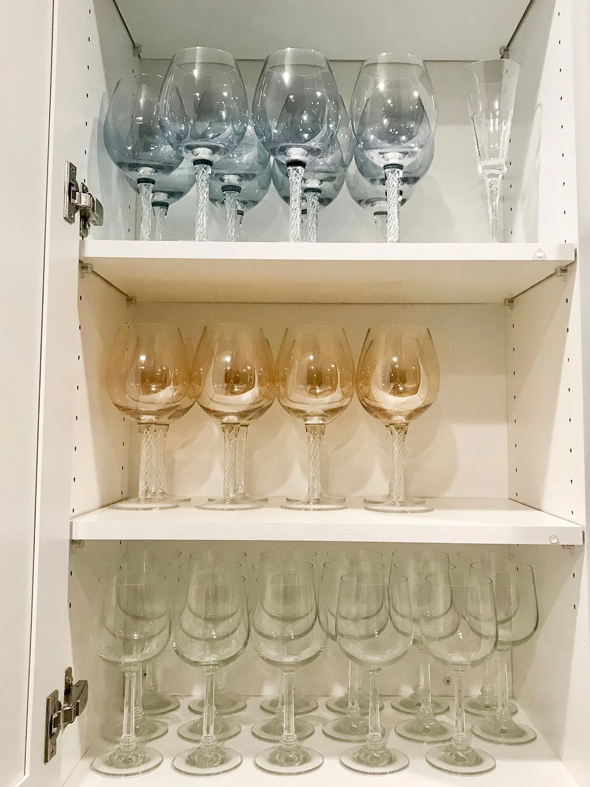 Cabinet with three shelves holding wine glasses of different colors and sizes, including blue, rose, and clear glasses, all upside down.