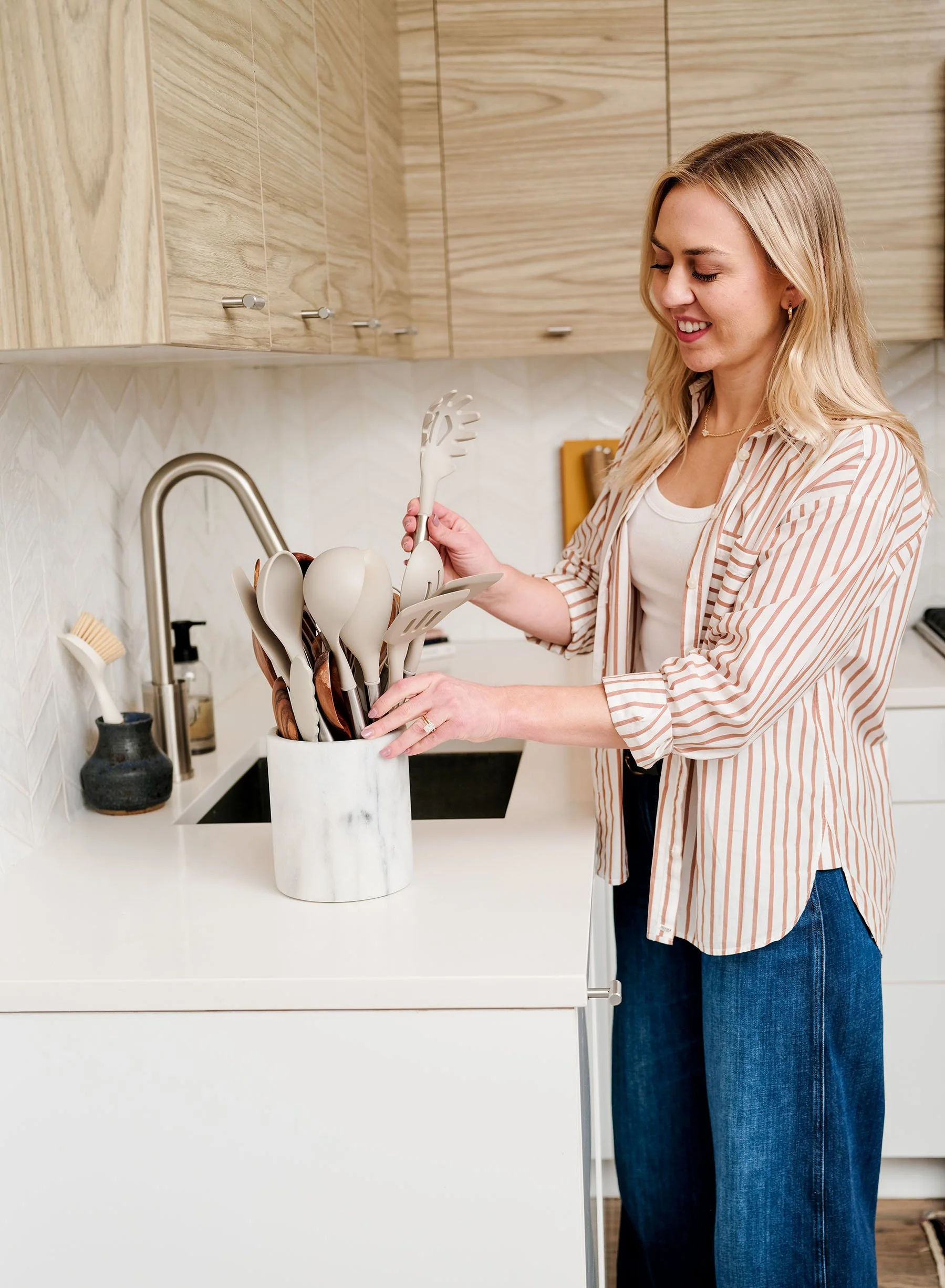 A woman with blonde hair, wearing a striped blazer and blue jeans, placing kitchen utensils into a white container on a kitchen countertop.