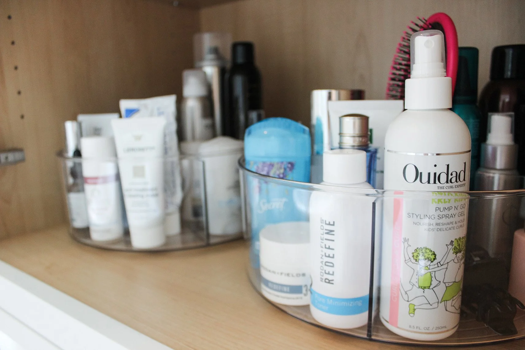 A wooden cabinet shelf with a clear organizer holding various personal care products, including bottles of hair styling spray, skincare items, and a hairbrush with pink bristles.