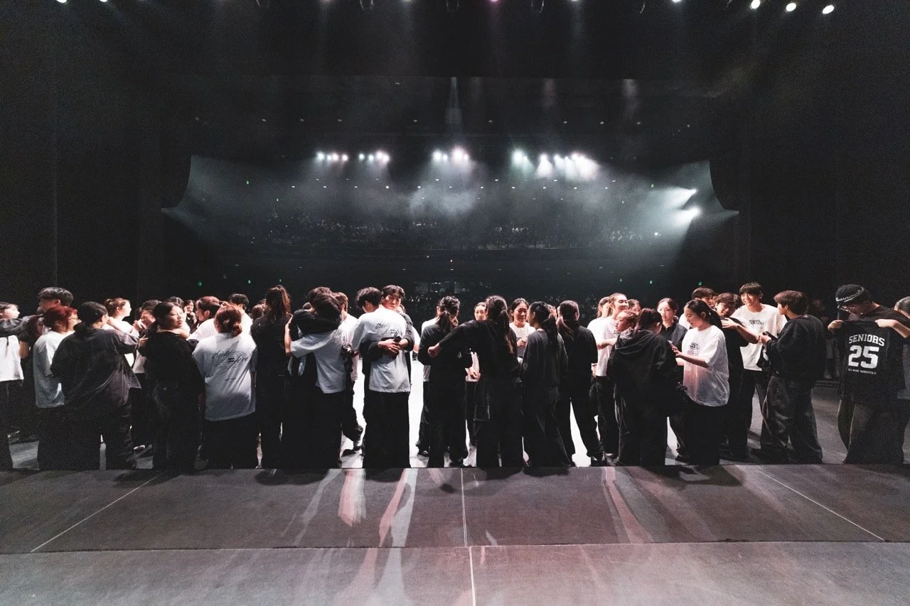 Group of people gathering backstage, preparing to go on stage for a performance, with stage lights shining in the background. Kids are dancing on stage in La Mirada, CA for their 34th annual concert, worshipping Jesus through singing and dance.