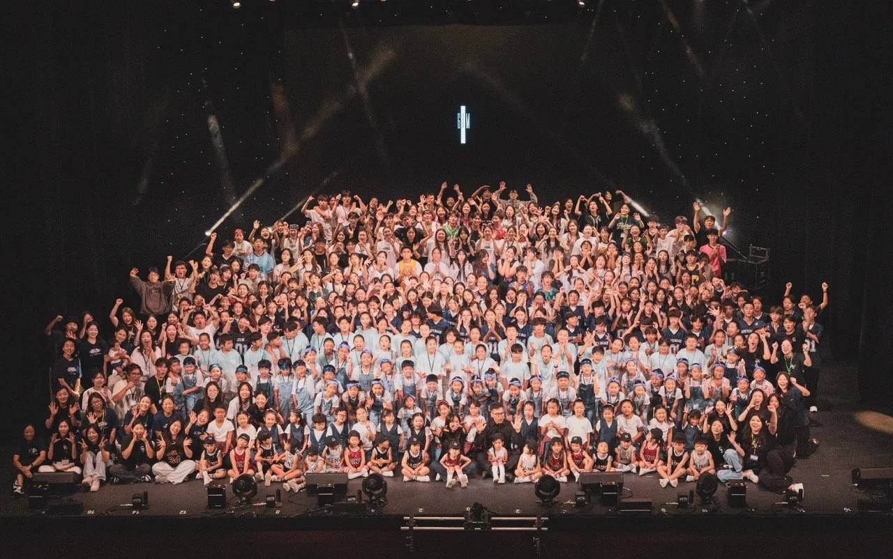 Large group of children and adults on stage celebrating, some raising their fists, with stage lights and a black background. Kids are dancing on stage in La Mirada, CA for their 34th annual concert, worshipping Jesus through singing and dance.