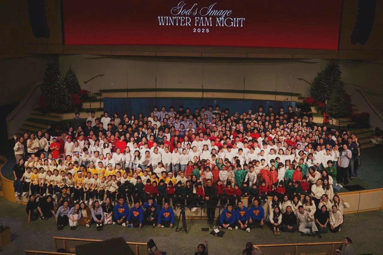 A large group of children and adults on stage for a Christmas performance, with Christmas trees and poinsettias, in front of a red screen that reads 'God's Image Winter Fam Night 2025'.