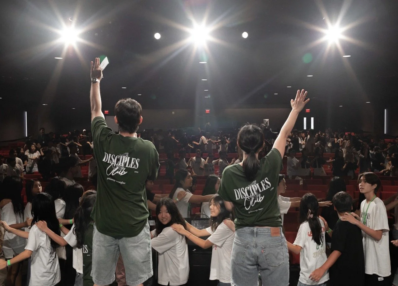 A large group of young people participating in a church or youth group event in a dark auditorium, two leaders leading them in worship or prayer with hands raised, and many children and teens standing in lines.