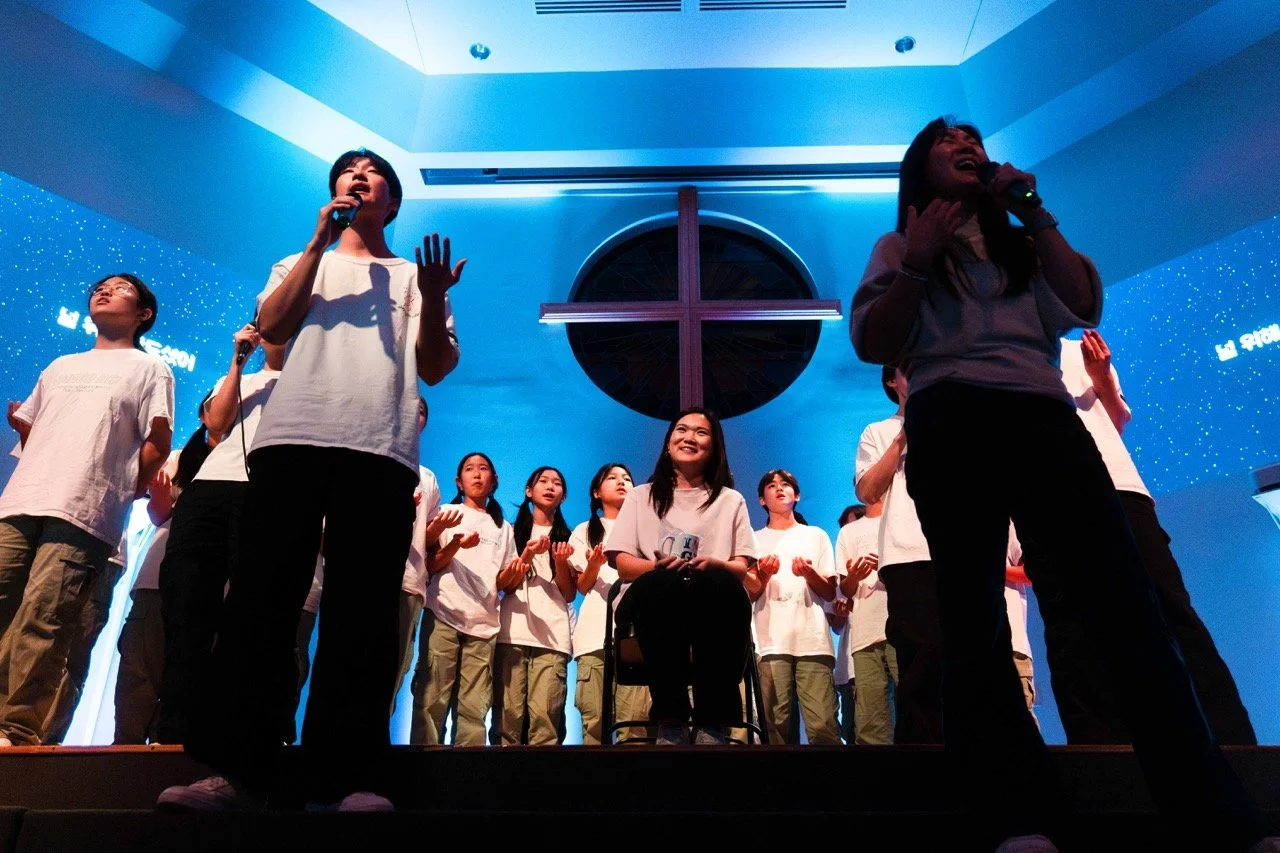 Group of young people on stage singing or performing in a church or religious venue with a cross in the background.