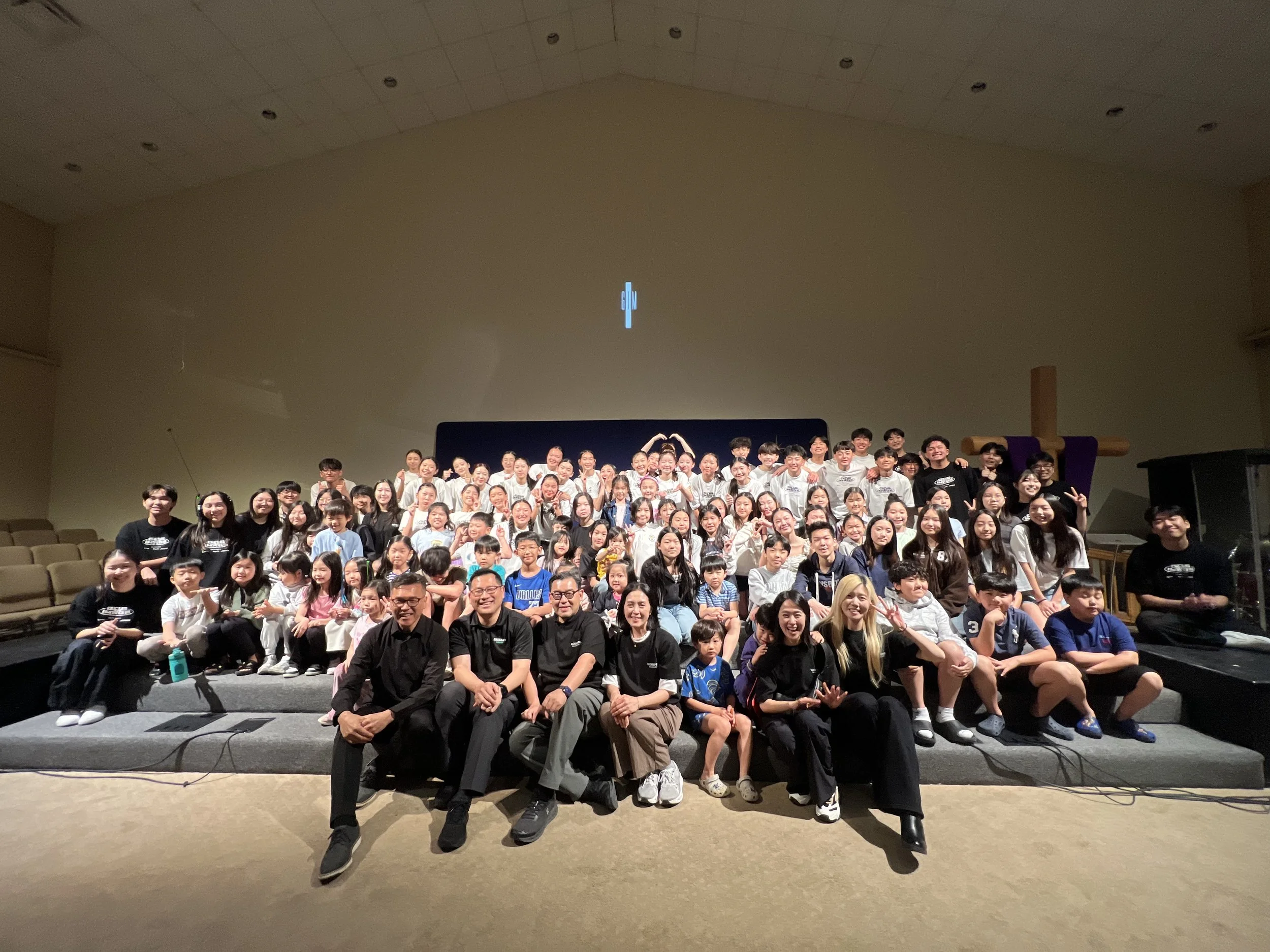 A large group of people, including children and adults, gathering on stage in a church or auditorium with a cross in the background, smiling for a group photo. Kids are dancing on stage in La Mirada, CA for their 34th annual concert, worshipping Jesu