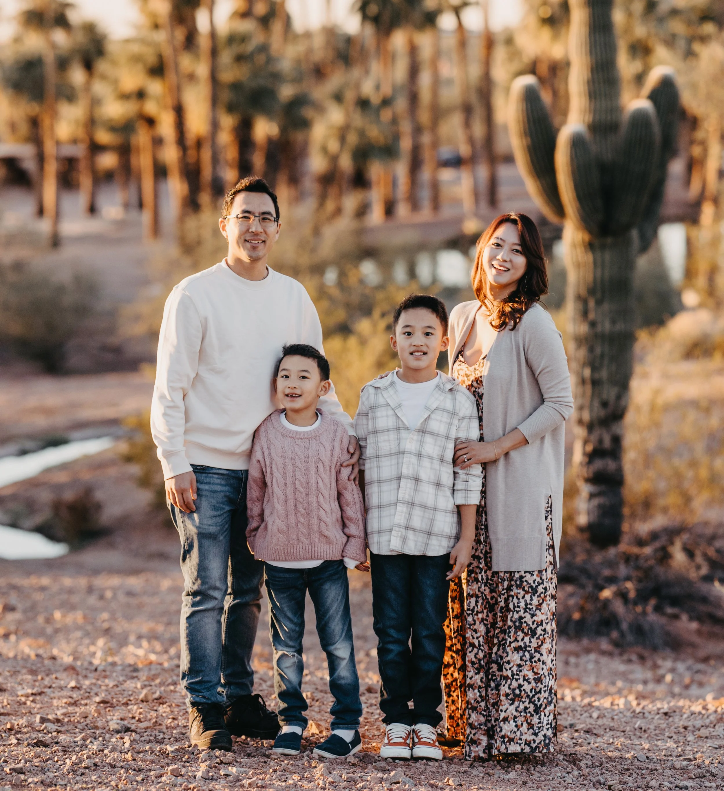 A family of five standing outdoors in a desert landscape during sunset. The family includes a man, a woman, and three children, with cacti and trees in the background.