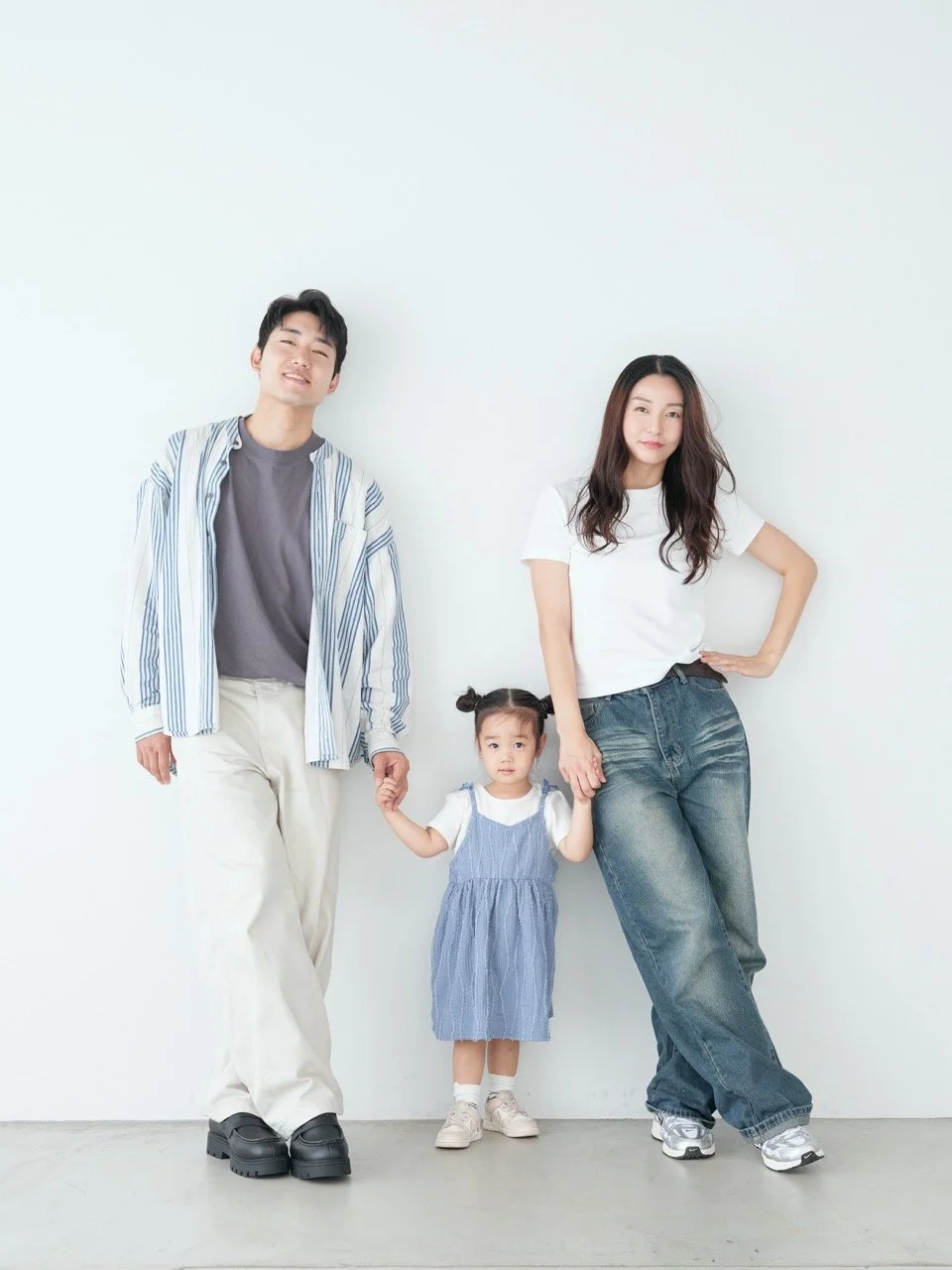 A family of three, an Asian man, woman, and young girl, standing against a white wall. The man and girl are holding hands, and the woman is standing with her hand on her hip. They are dressed casually with the girl in a blue dress and the adults in comfortable, stylish clothes.