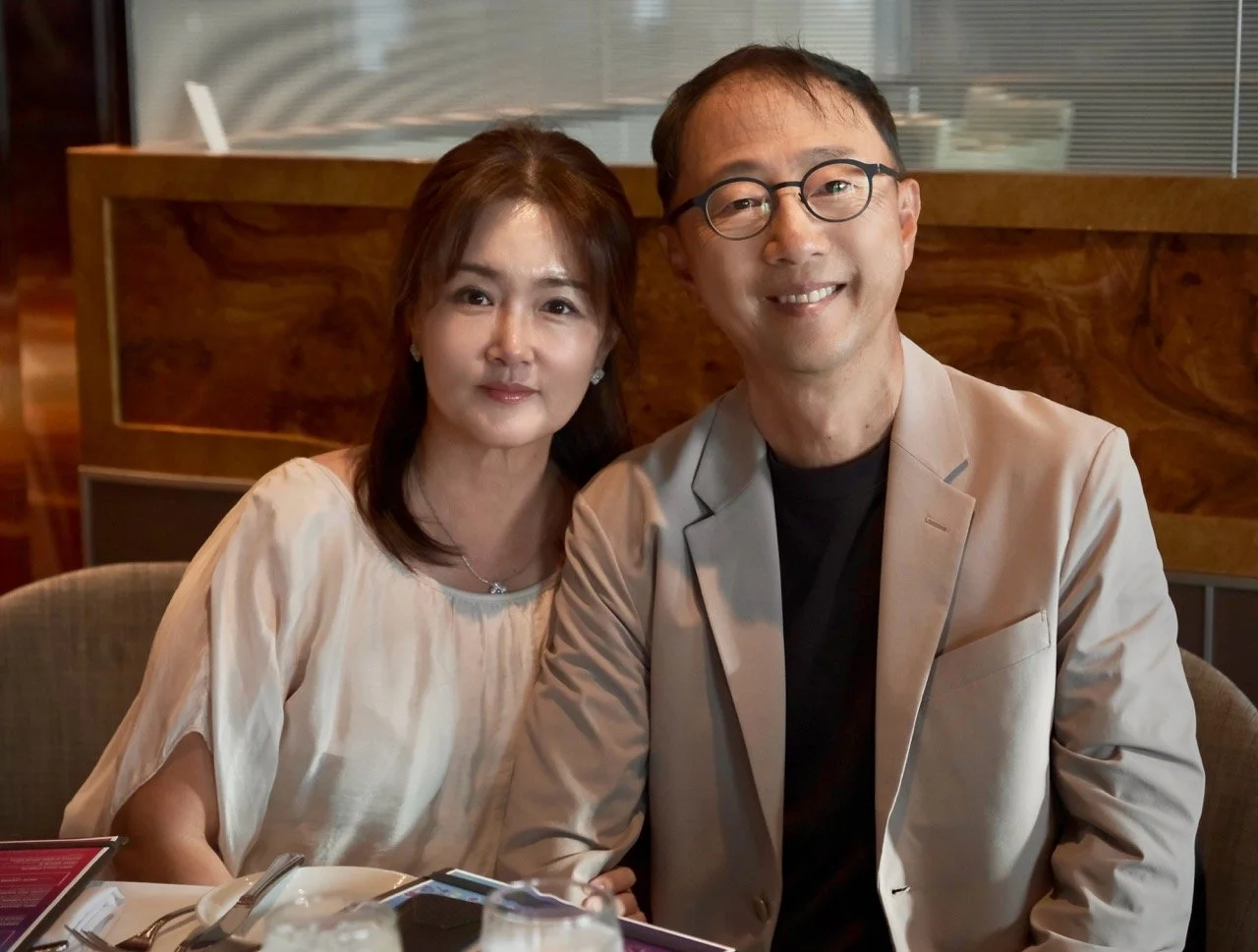 A man and woman sitting together at a restaurant, smiling at the camera. The woman is on the left wearing a cream-colored blouse, and the man is on the right wearing glasses and a light beige blazer.