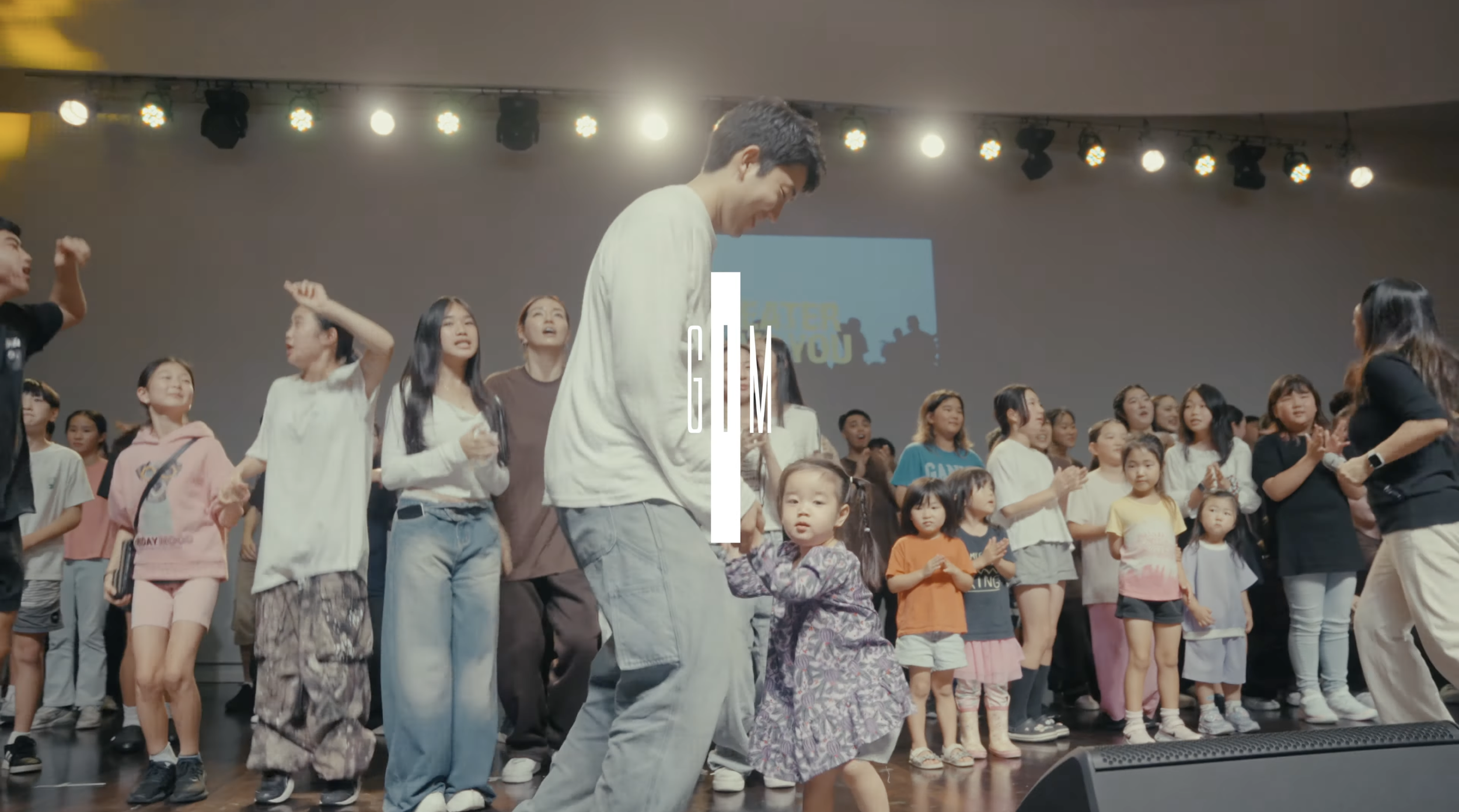 Group of children and adults on stage at a gym or event space, some dancing and others observing, with a large screen and stage lights in the background. Australia Tour 2025