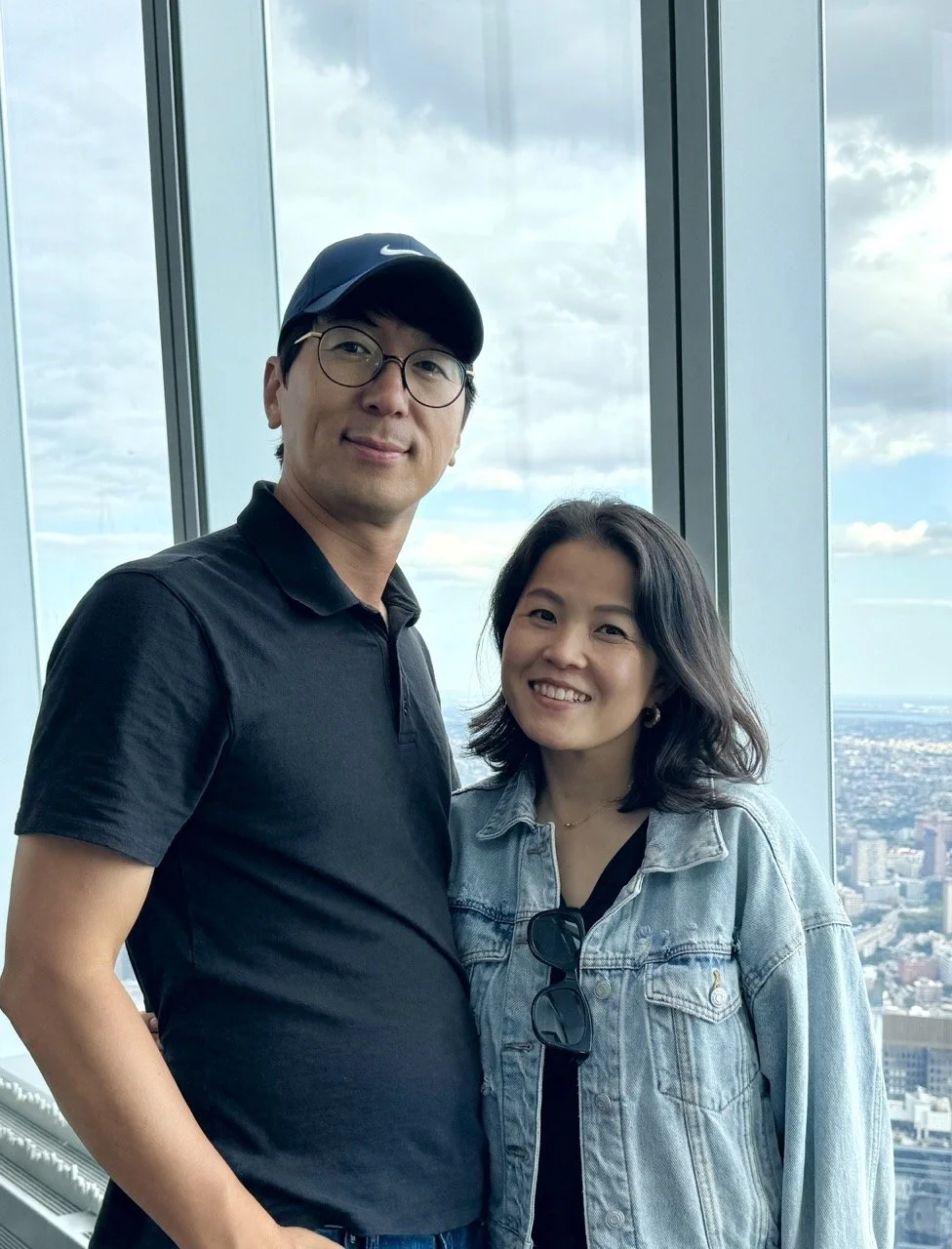 A man and woman standing close together in front of large windows with a city skyline in the background. The man is wearing a black Nike cap, glasses, and a black polo shirt. The woman is wearing a denim jacket, black top, and sunglasses hanging from her jacket. Both are smiling.