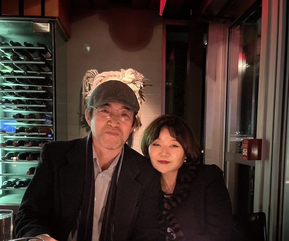 A man wearing glasses and a cap sitting next to a woman with short dark hair, at a restaurant or bar with wine bottles in a wine fridge in the background.
