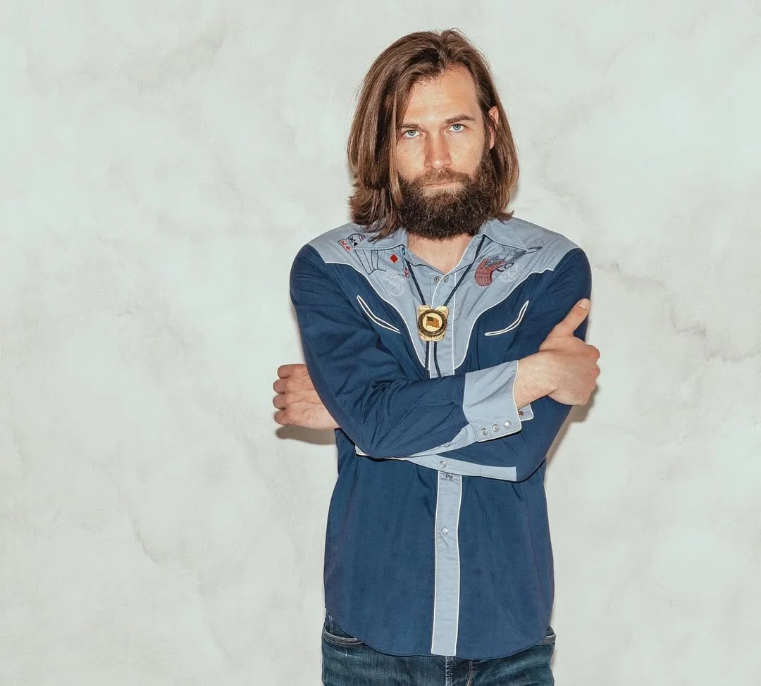 A man with long brown hair and a beard, wearing a blue western shirt with embroidery, standing with arms crossed and looking at the camera against a plain light-colored wall.