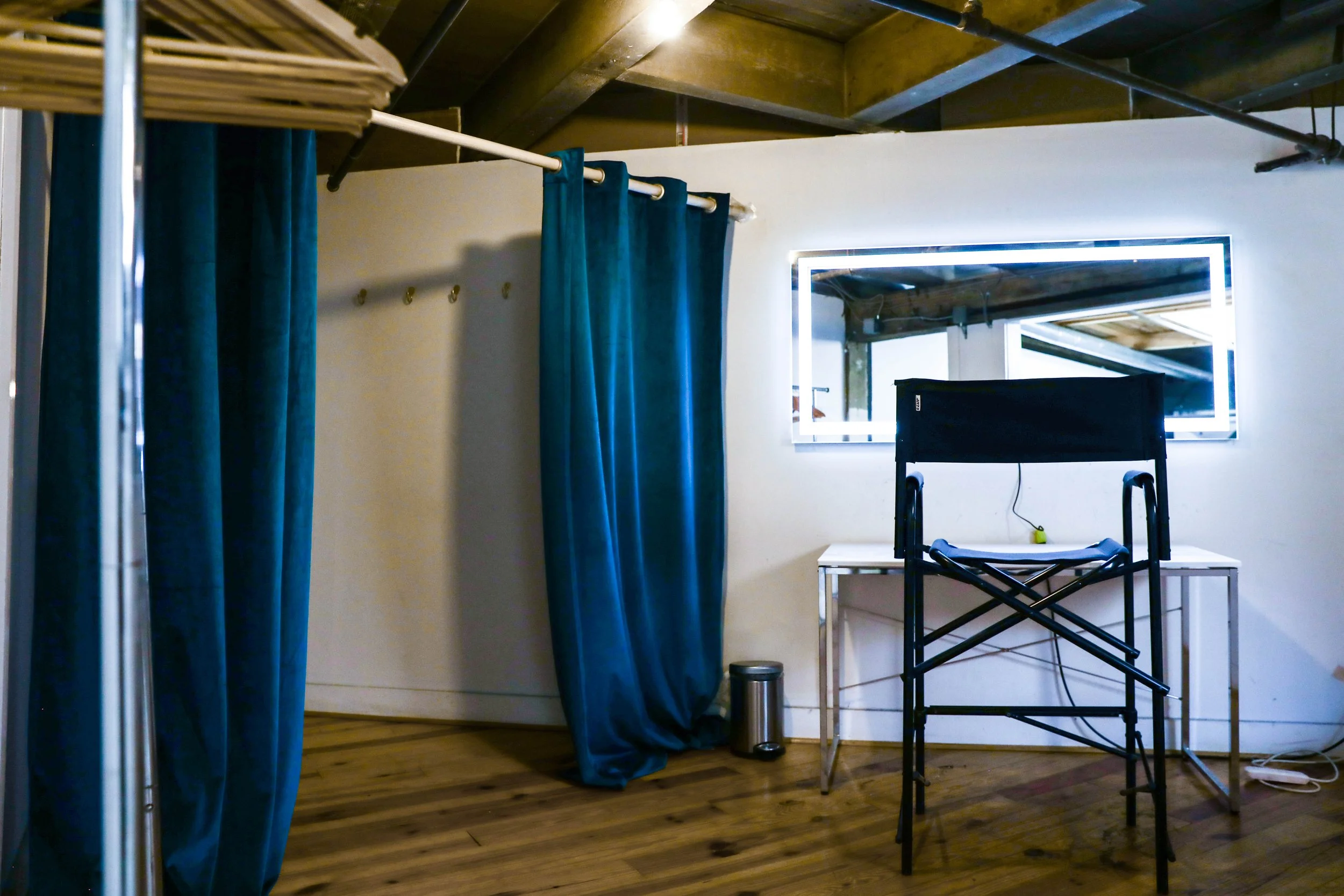 Room with wooden floor, white wall, and ceiling with exposed wooden beams. Blue curtains hanging on a curtain rod, desk with a folding chair, large illuminated mirror on the wall, small trash can, and some cables visible.