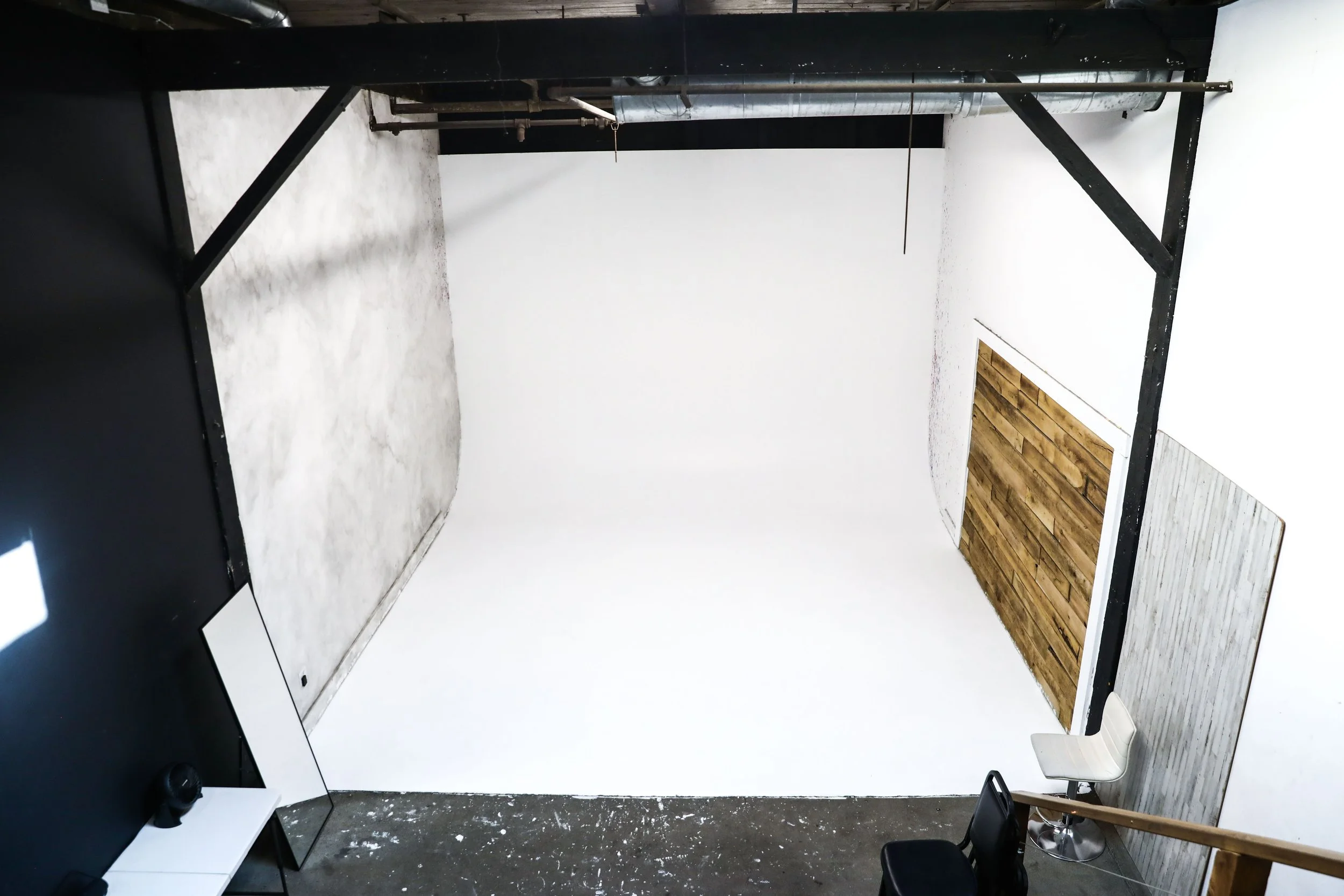 Photo of an indoor studio space with a white cyclorama wall, a small white table with a mirror and camera, a black chair, a white high chair, and a wooden panel leaning against a section of the wall. Exposed pipes and ductwork are visible on the ceil
