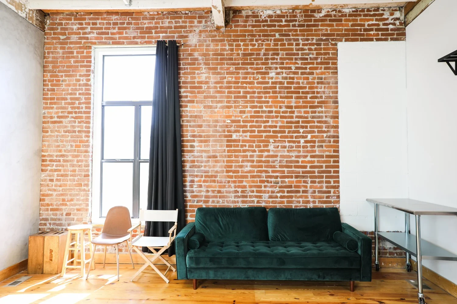 Living room with exposed brick wall, large window with black curtains, green velvet sofa, two chairs (one white with wooden arms and one beige), wooden crate, and metal rolling cart, with sunlight streaming onto a wooden floor.