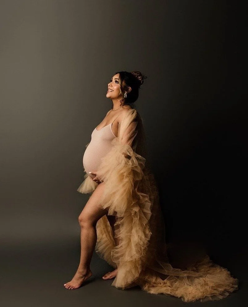 A pregnant woman in a beige dress with sheer, ruffled sleeves, standing barefoot against a dark background, smiling and looking upward.