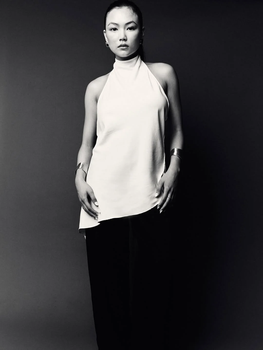 A black-and-white photo of a woman with makeup and short hair, wearing a sleeveless high-neck top and black pants, standing against a plain dark background.