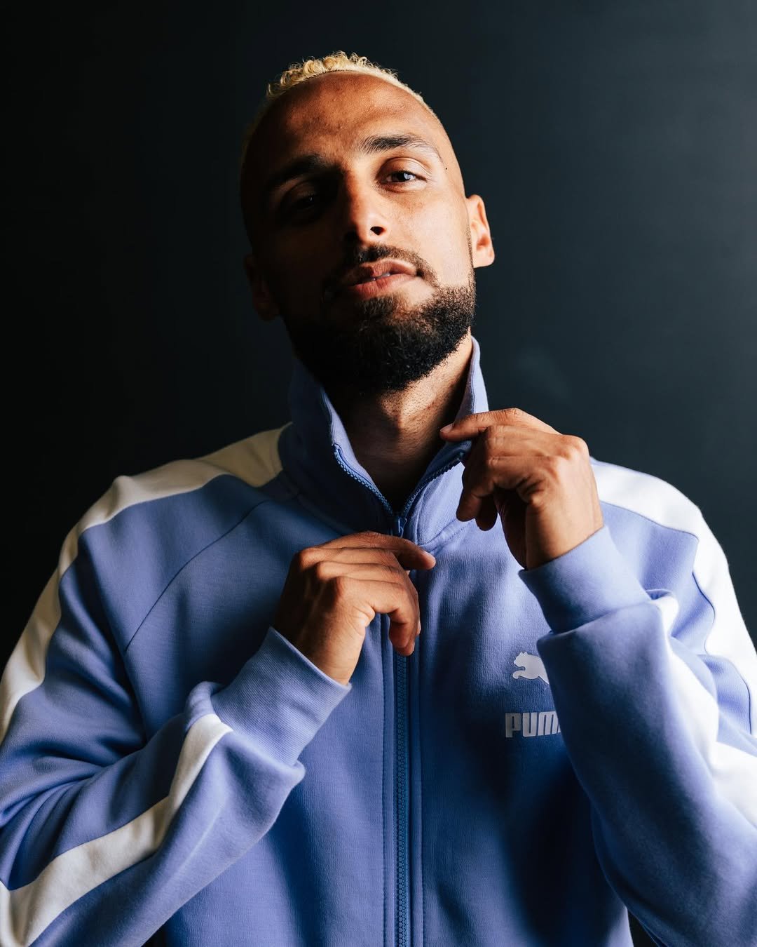 Man adjusting his blue Puma sports jacket against a dark background.