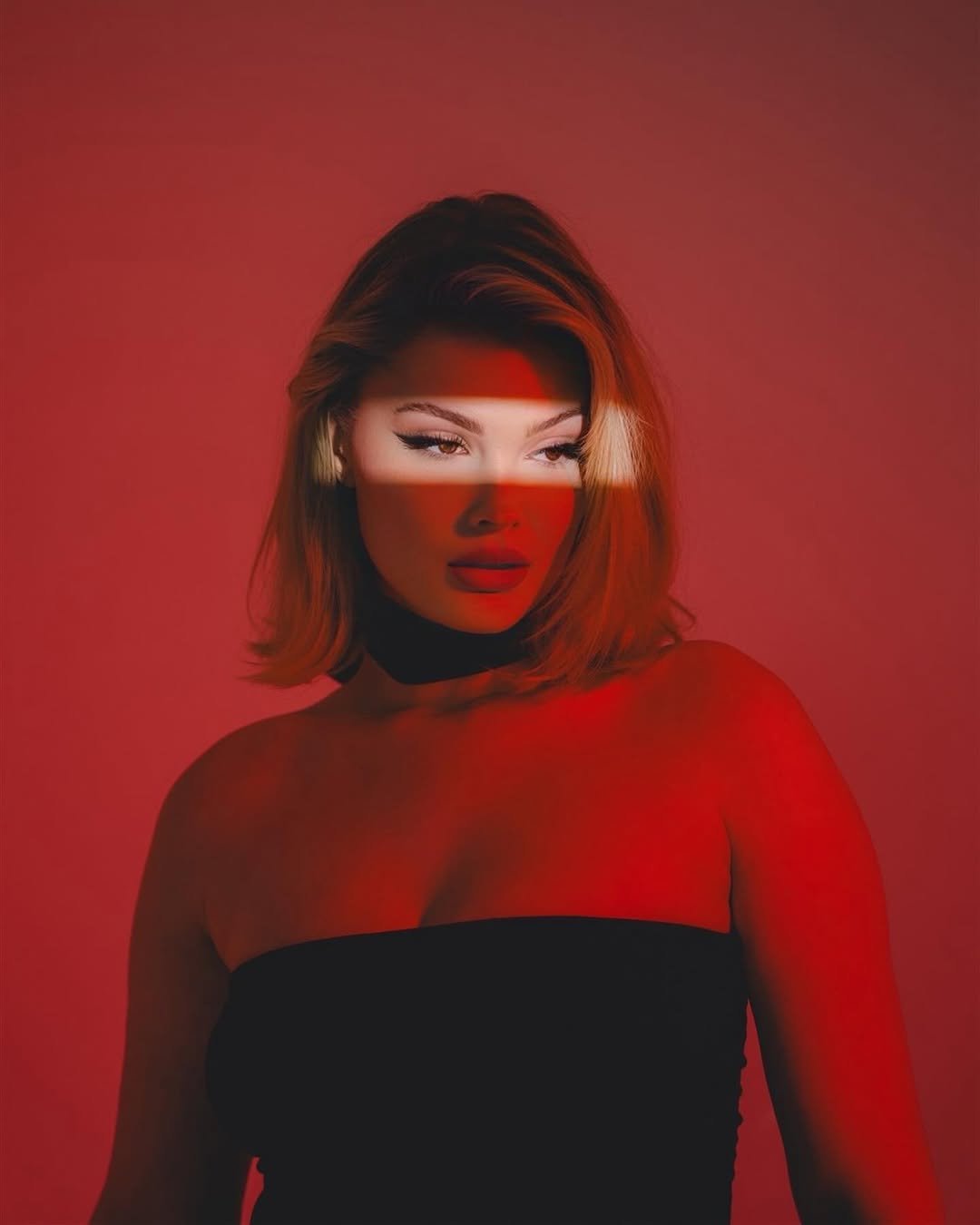 A woman with blonde hair, wearing a black strapless top, stands against a red background with a horizontal light stripe across her face.