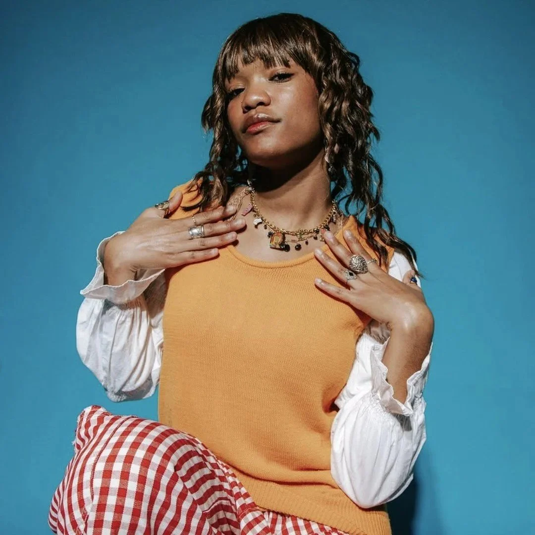 Young woman with curly hair wearing a mustard yellow top, white puffed sleeves, and red checkered pants, posing against a blue background with jewelry.