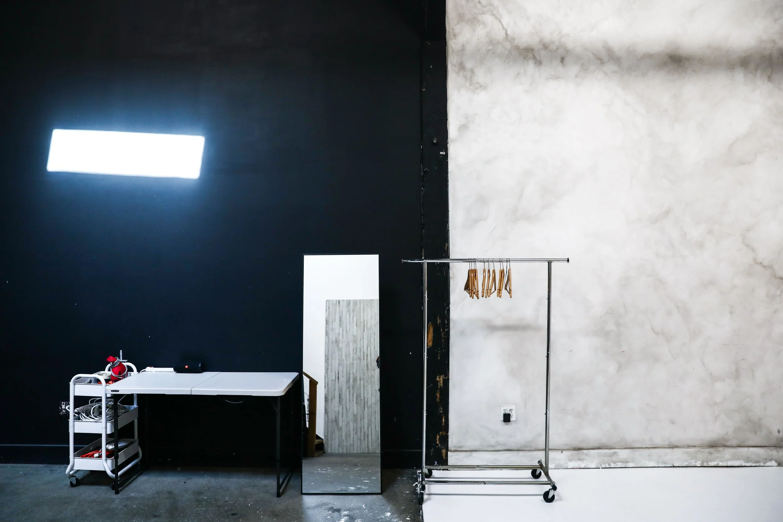 Minimalist interior with a black wall on the left and a white wall on the right. There is a white table, a small rolling cart with shelves and cords, a large mirror leaning against the wall, and a metal clothing rack with wooden hangers. A doorway is