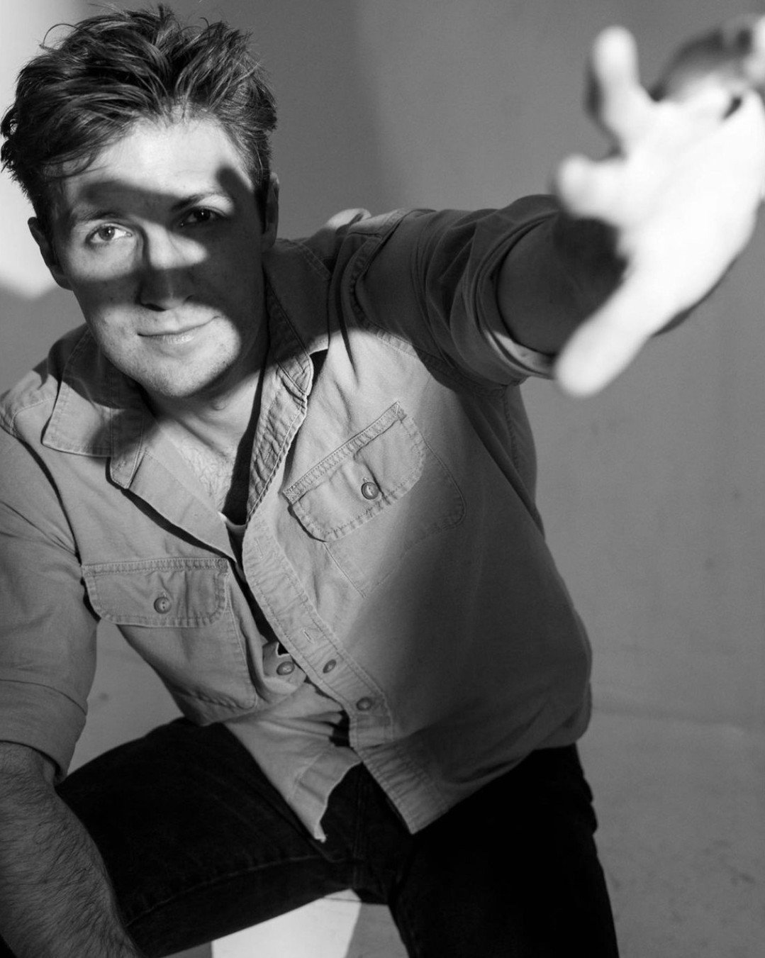 A young man with tousled hair wearing a button-up shirt reaching out toward the camera, with a shadow across his face in a black and white photo.