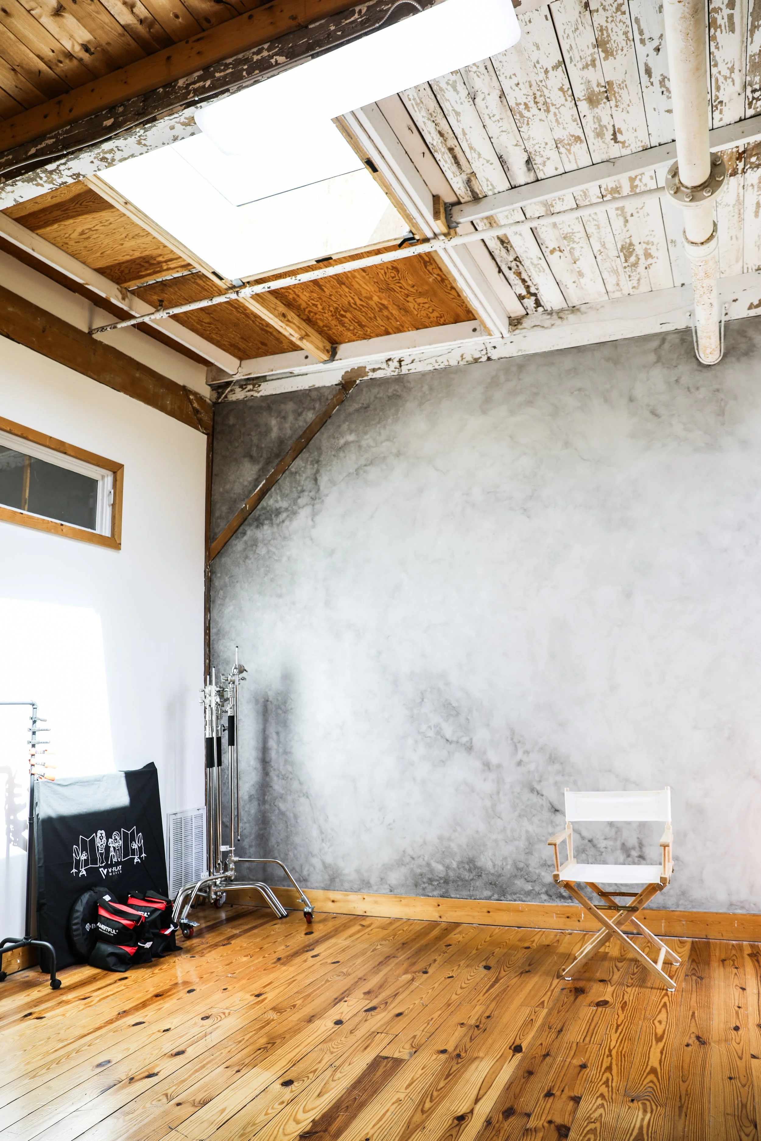 An empty art studio with a wooden floor, a textured grey wall, a director's chair, photography equipment, and a ceiling with exposed ceiling tiles and pipes.