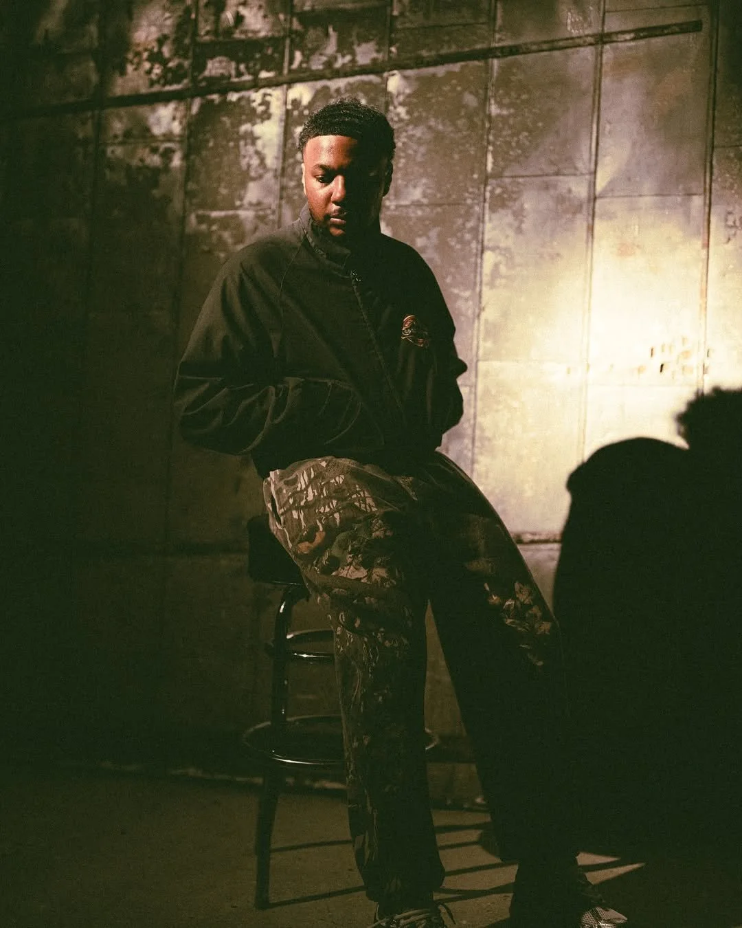 A young man sits on a high stool against a grimy, textured wall with peeling paint, in a dimly lit, industrial-style setting.