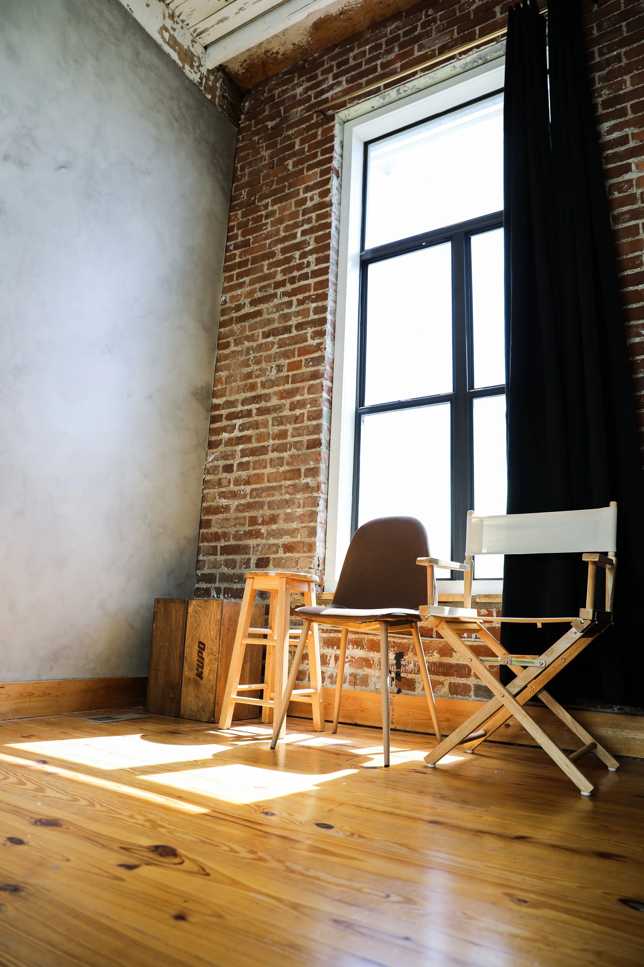 Two chairs, one brown and one white, appear to be in an industrial-style room with exposed brick wall, large window, and hardwood floor, with sunlight streaming in.